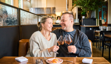 A couple sits closely at a restaurant table, smiling and clinking glasses of rosé wine, with a dessert dish in front of them.