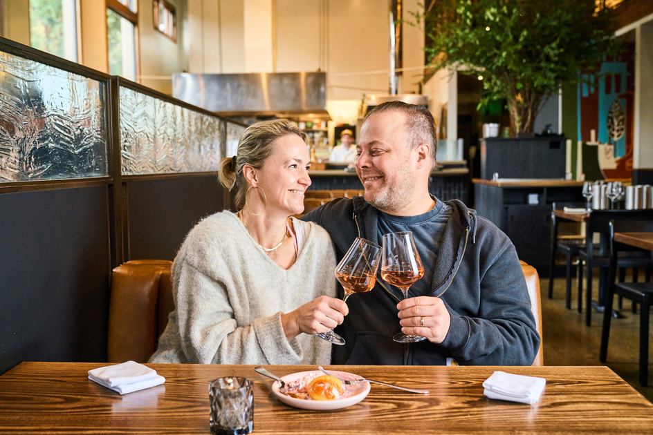 A couple sits closely at a restaurant table, smiling and clinking glasses of rosé wine, with a dessert dish in front of them.