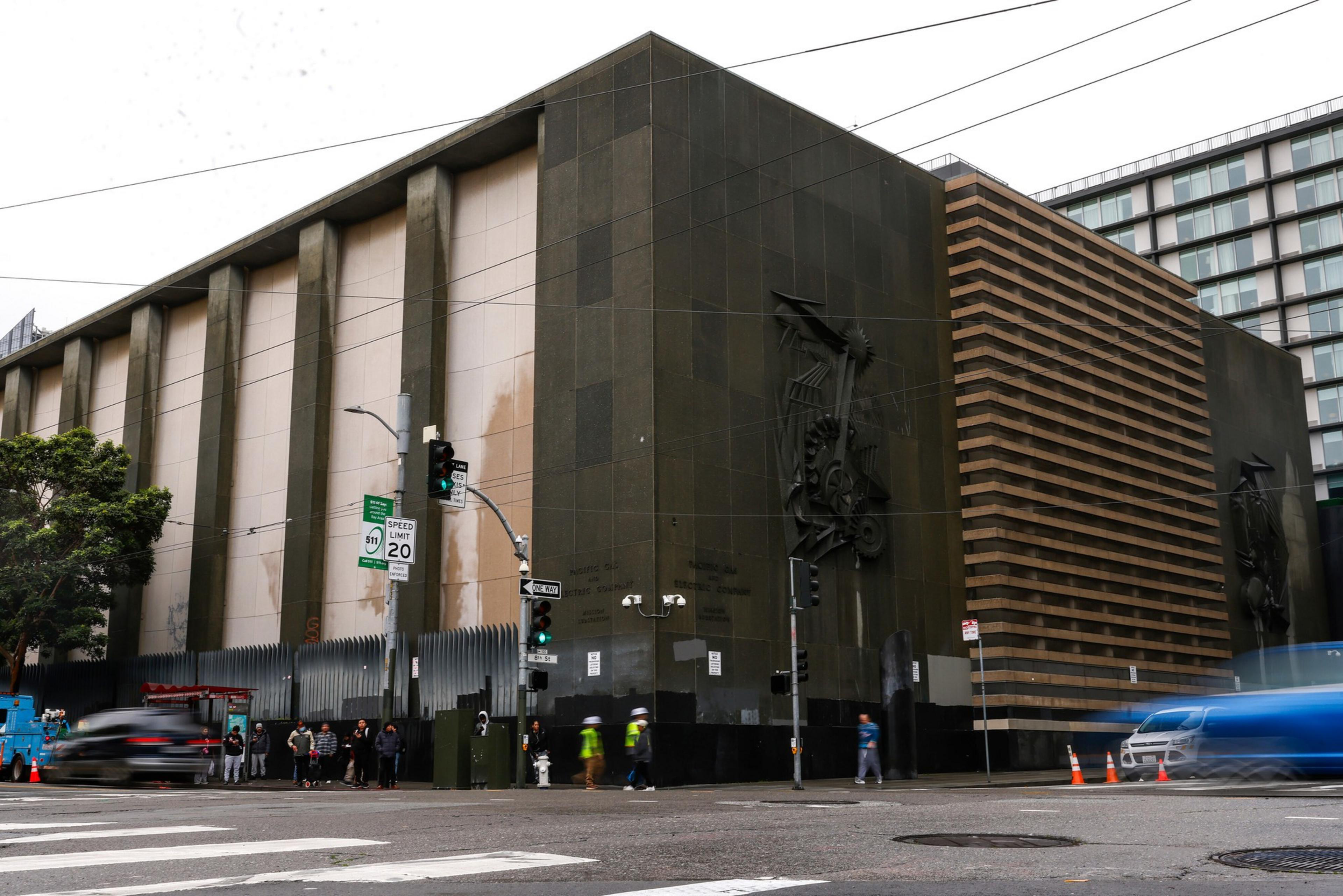A large, dark, blocky building with vertical columns and abstract metal sculptures on its walls sits at a busy urban intersection with pedestrians and cars.