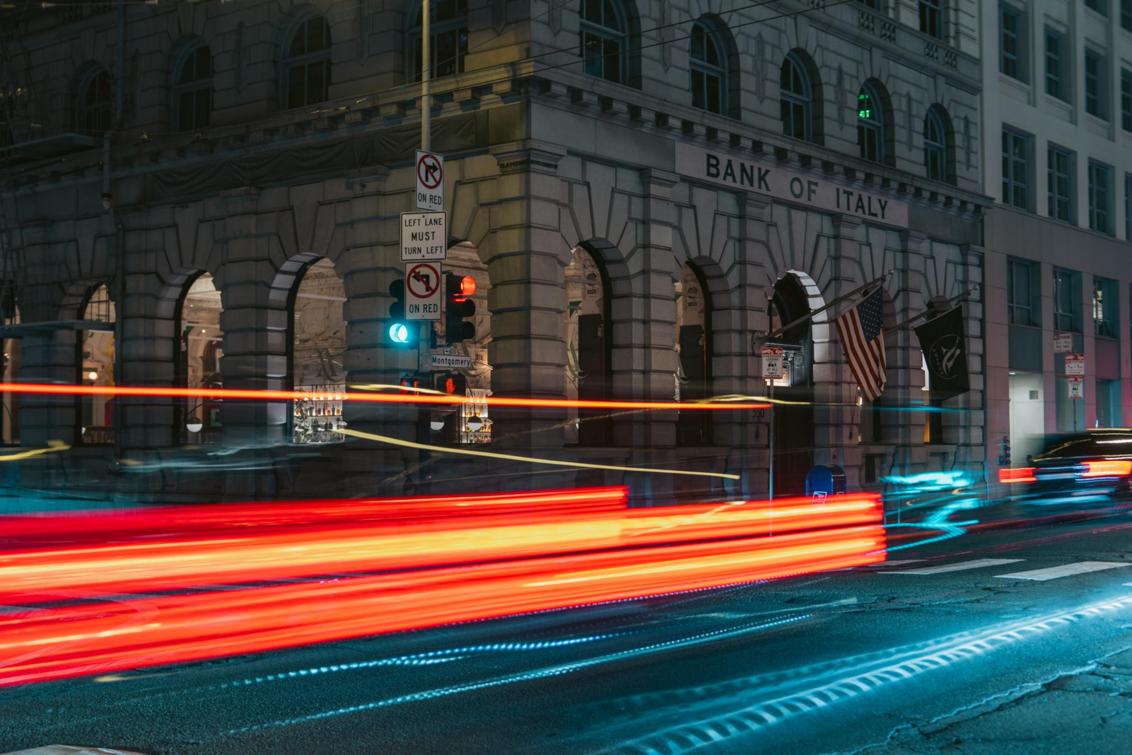 A street corner at night shows a Bank of Italy building with light trails from passing cars and traffic signals illuminated.