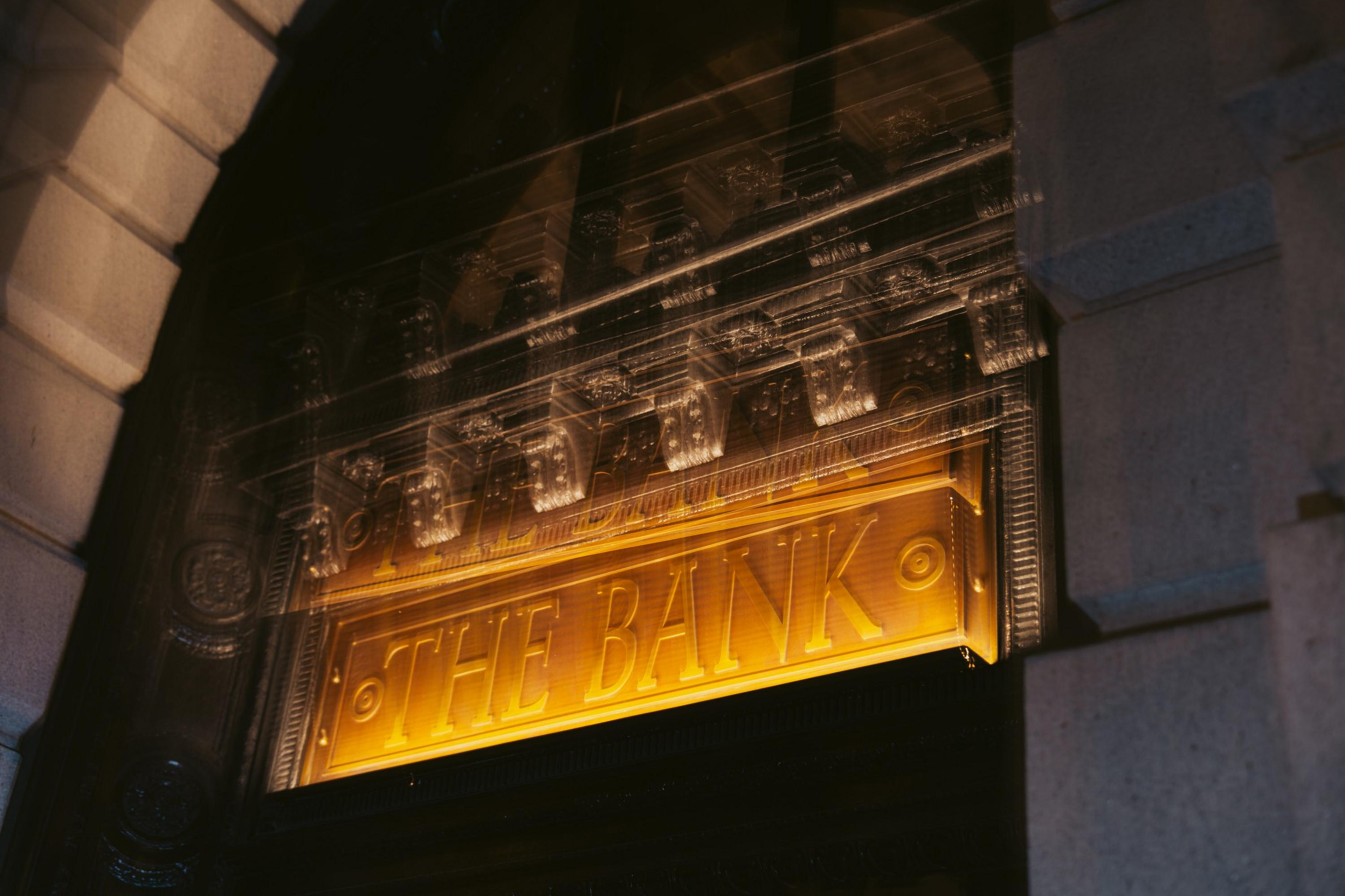 A golden sign reading “THE BANK” is mounted above a dark entrance, with ornate architectural details and a blurred double exposure effect.
