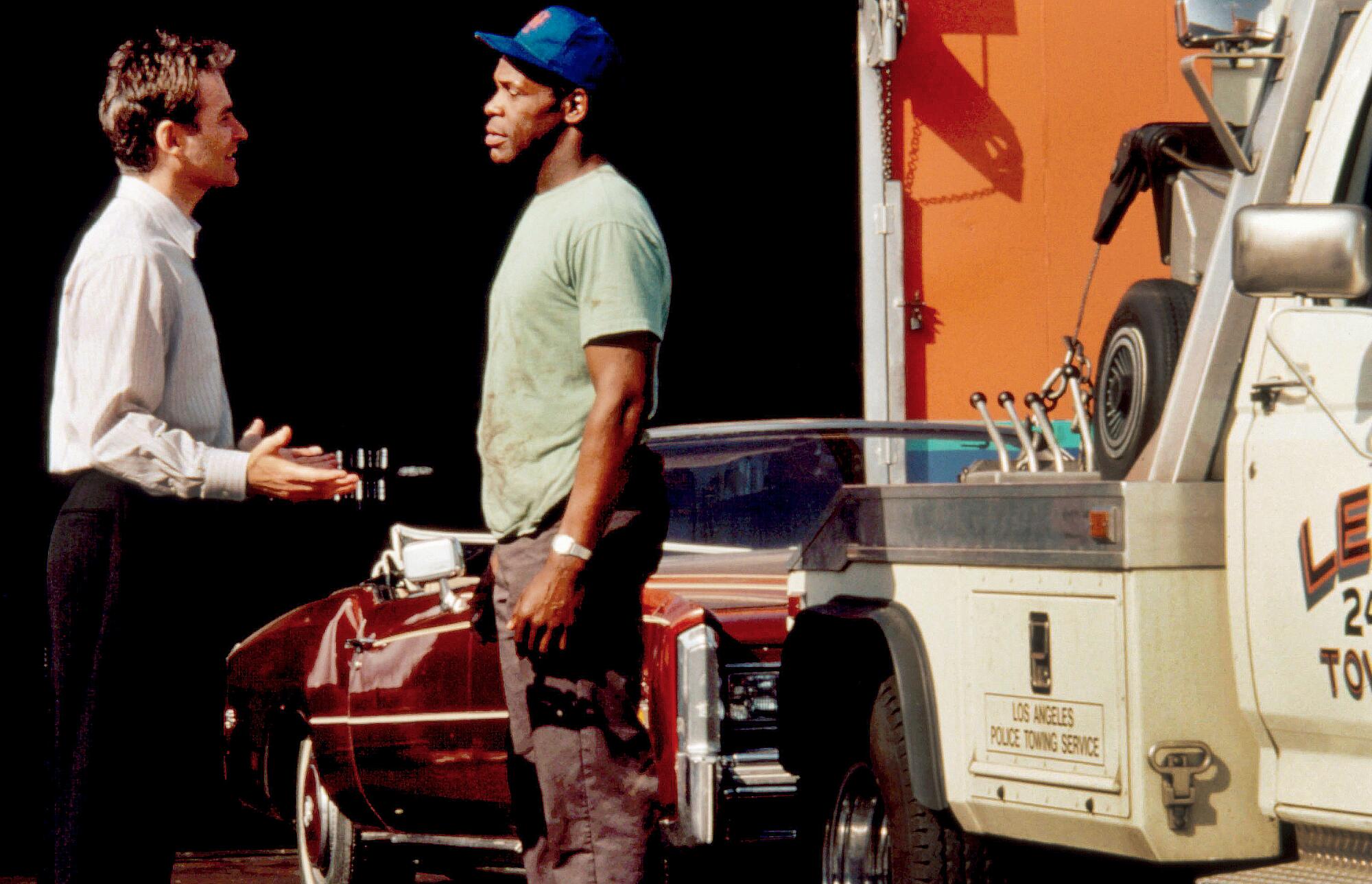 Two men have a discussion in front of a red convertible getting towed.