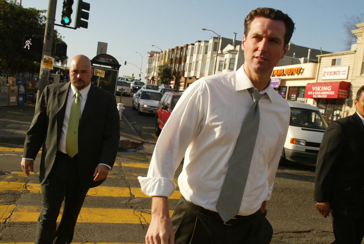 Gavin Newsom in a white dress shirt and tie walks down a sidewalk