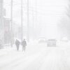 Two individuals walk along a street; they, the vehicles behind them, and the building next to them are blurred by snowfall.