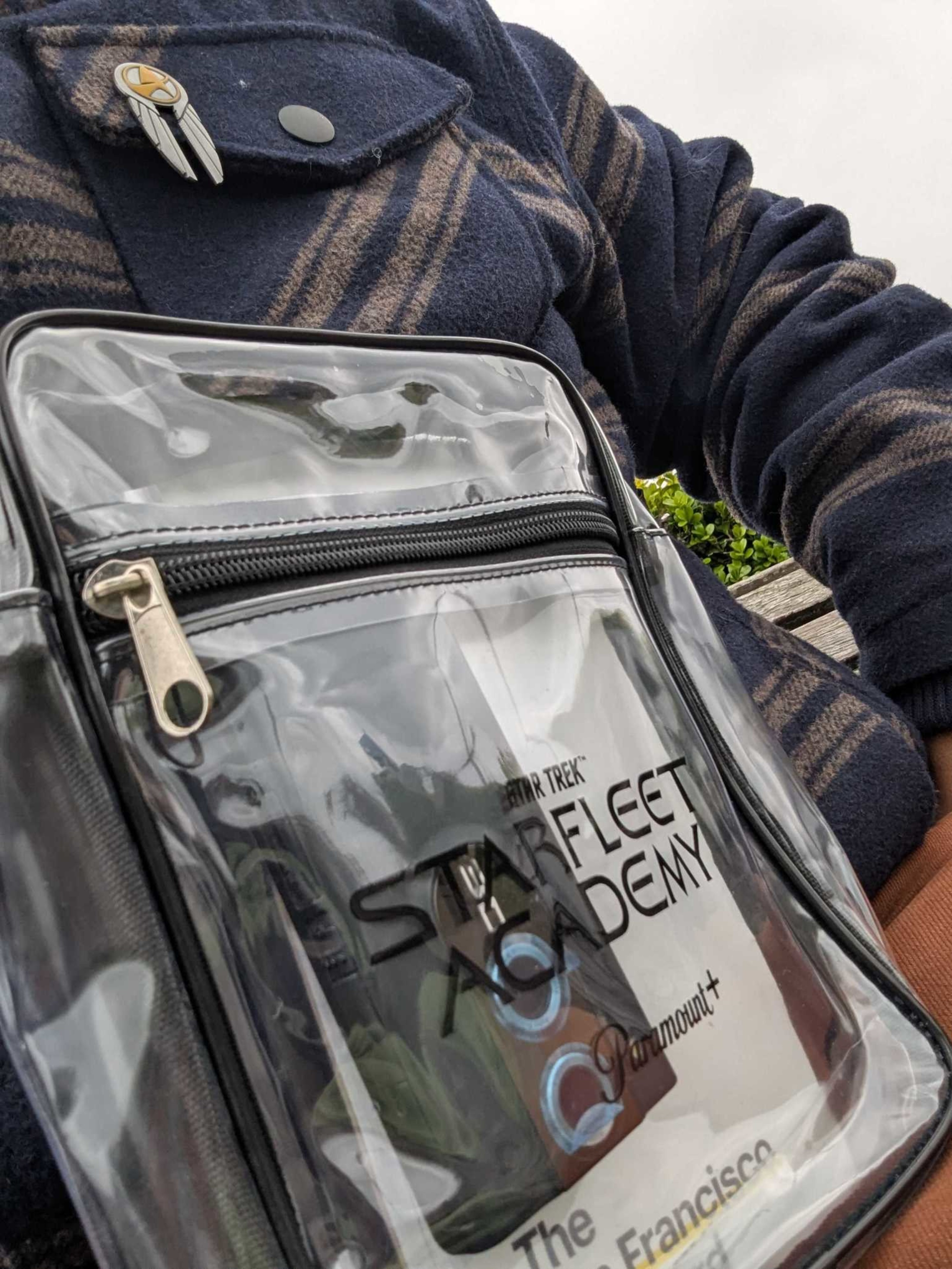 A person wearing a blue and brown striped jacket with a Star Trek pin holds a clear bag labeled “STARFLEET ACADEMY Paramount+ The San Francisco.”