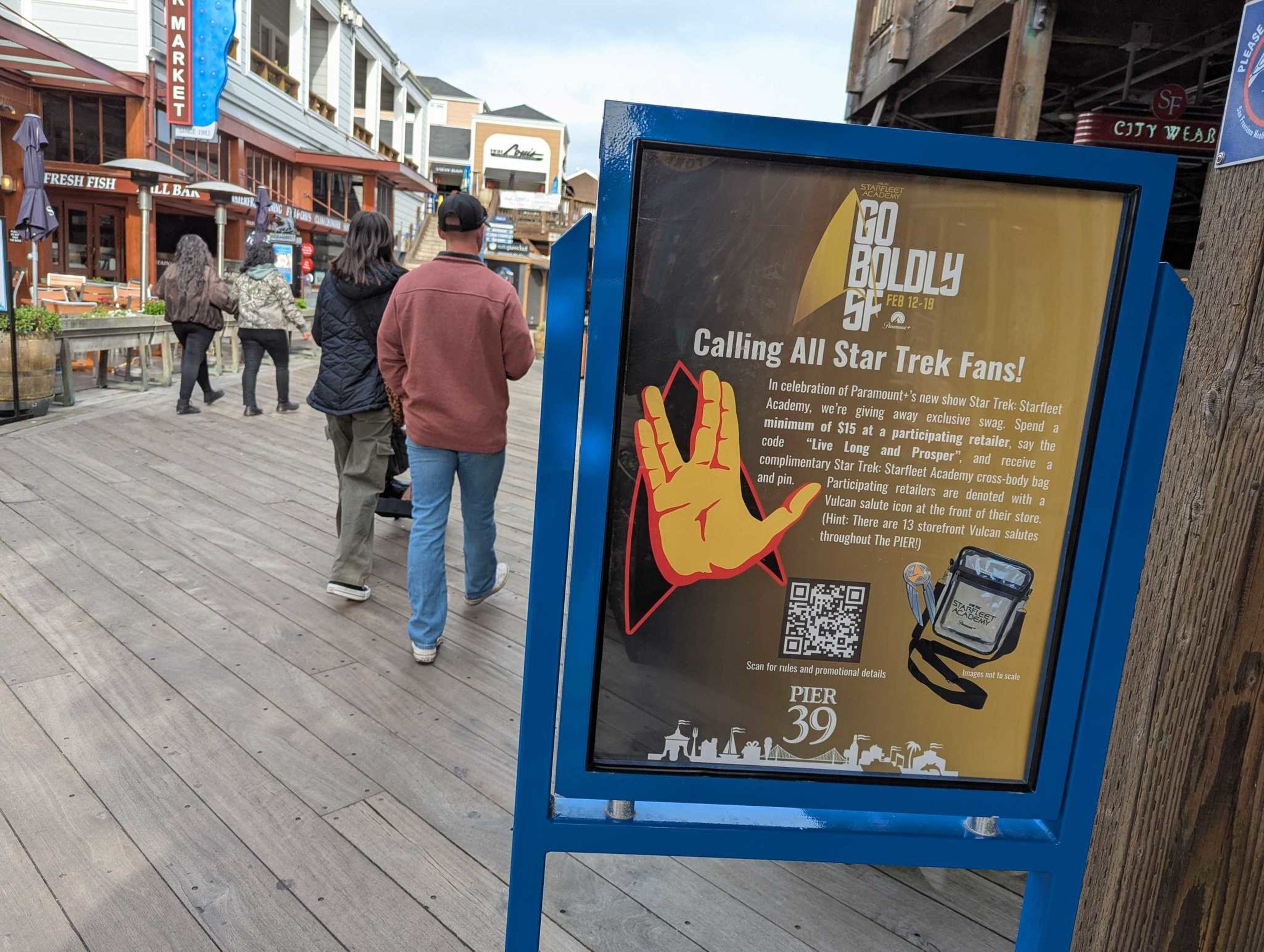 A blue sign at Pier 39 invites Star Trek fans to spend $15 for exclusive swag, featuring a red Vulcan salute hand and a cross-body bag image.