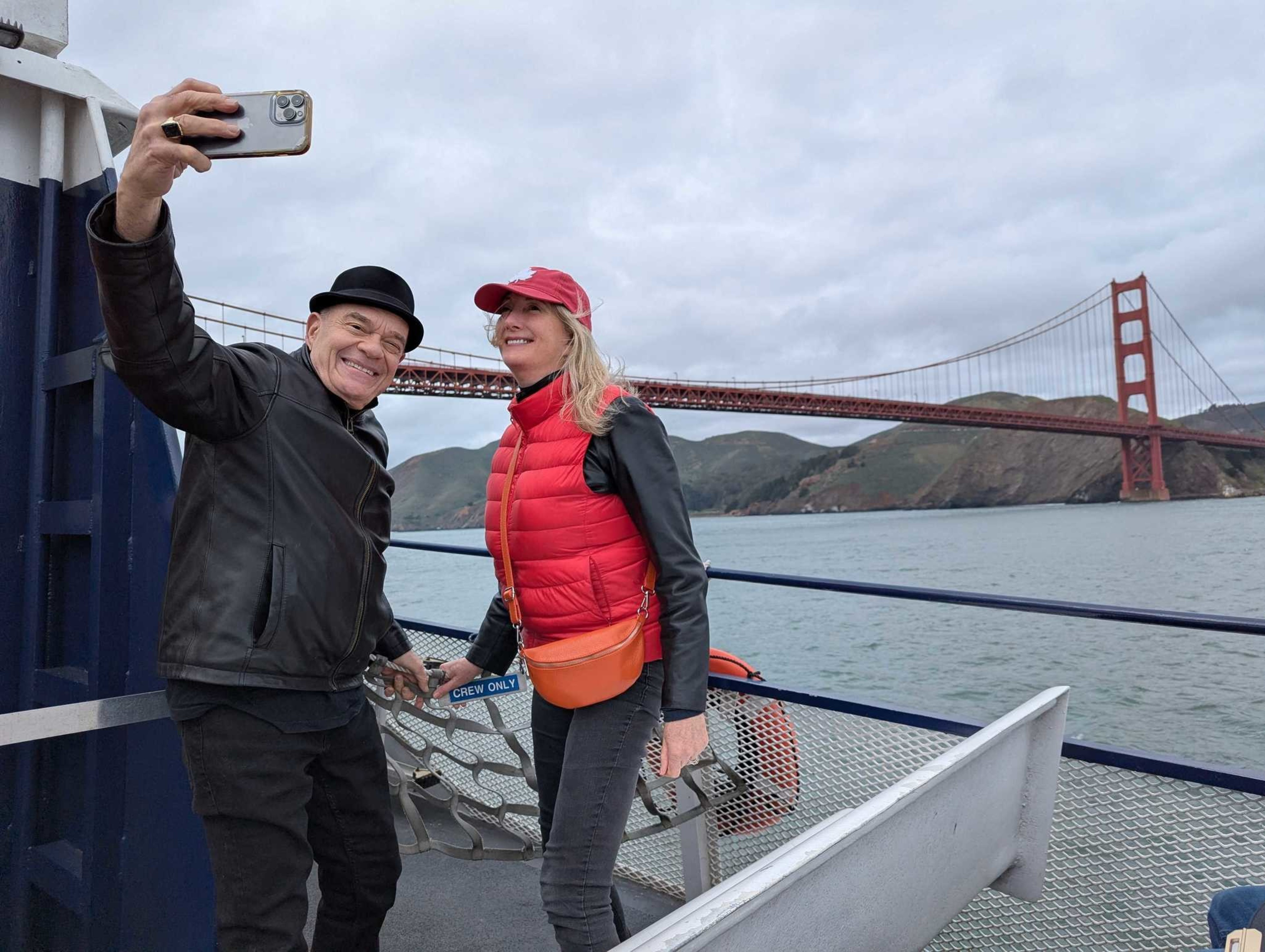 A man in a black jacket and hat takes a selfie with a smiling woman in a red vest and cap on a boat, with the Golden Gate Bridge in the background.