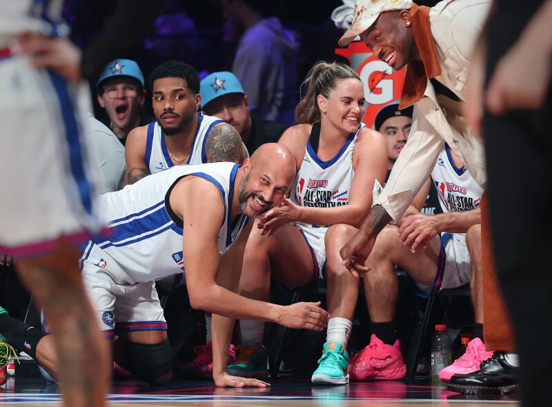 Celebrity Keegan-Michael Key kneels and laughs with teammates sitting on a bench during a game.