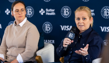 Two women sit in armchairs against a backdrop with logos, one speaks into a microphone while gesturing, the other listens holding a microphone in her lap.