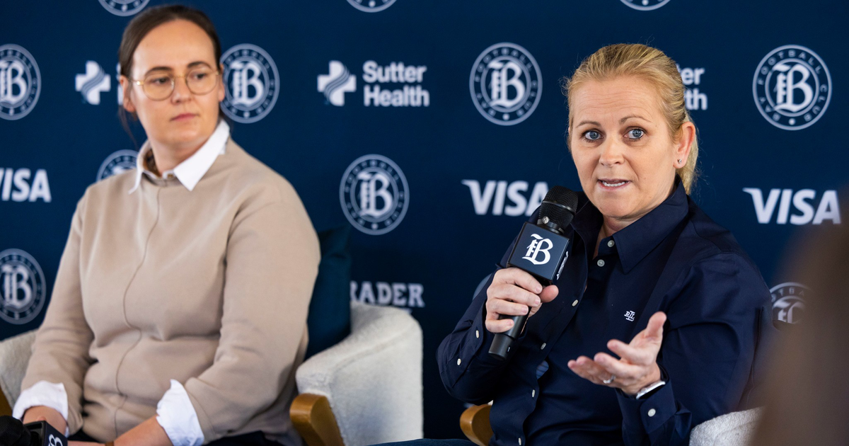 Two women sit in armchairs against a backdrop with logos, one speaks into a microphone while gesturing, the other listens holding a microphone in her lap.
