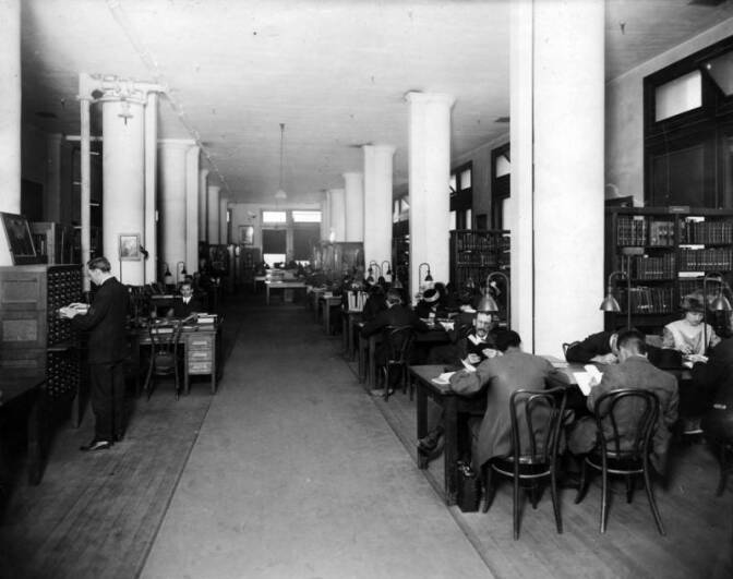 A black and white photo shows a room with pillars and desks. People sit and read with bookshelves lining one wall.