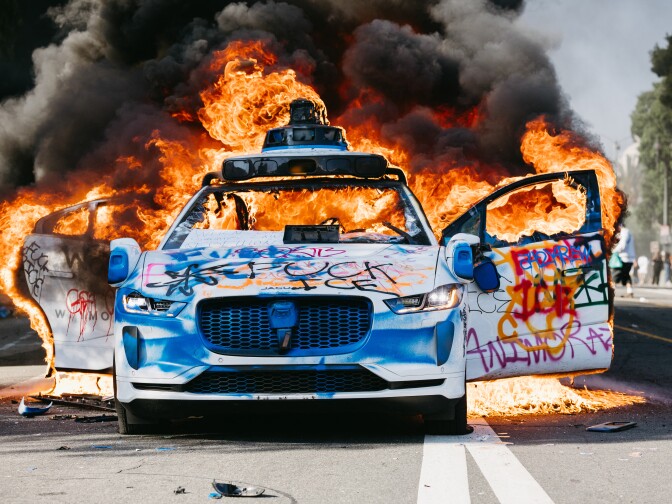 A Waymo car burns in the street as smoke billows during protests in Los Angeles on Sunday. Following a series of aggressive federal immigration operations in the city, tensions escalated when ICE agents conducted a high-profile raid on a Home Depot location.