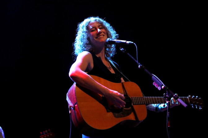 A light-skinned woman playing acoustic guitar onstage.