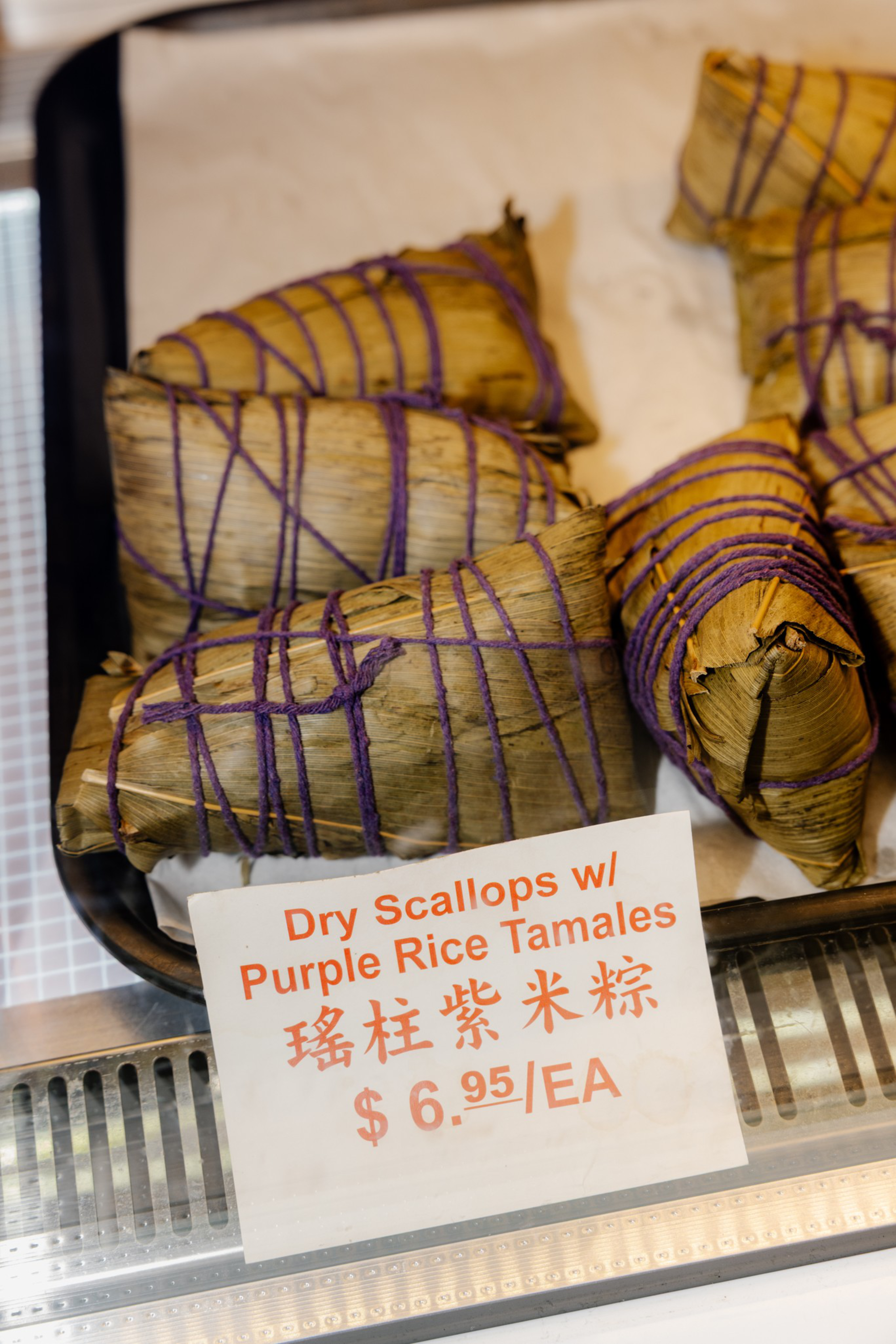Triangular tamales wrapped in dried leaves and tied with purple string, labeled as Dry Scallops with Purple Rice Tamales, priced at $6.95 each.