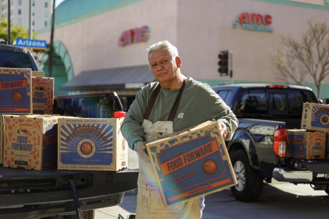 A portrait of Eleu, who's a man with a medium skin tone and gray hair. He's smiling at the camera while loading a box of Food Forward produce into a truck.