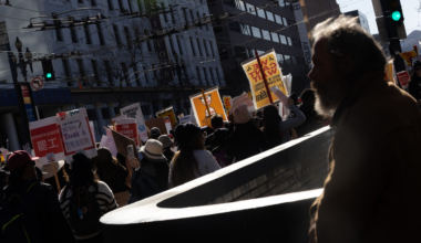 A crowd of people holds protest signs in a city street while a bearded man watches from the side, with strong sunlight casting shadows.