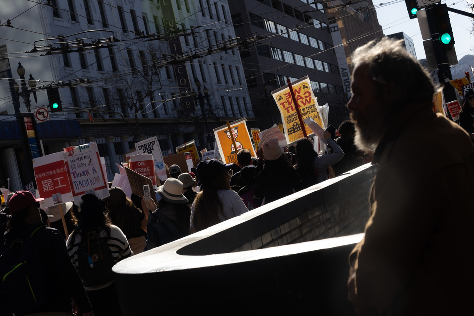 A crowd of people holds protest signs in a city street while a bearded man watches from the side, with strong sunlight casting shadows.