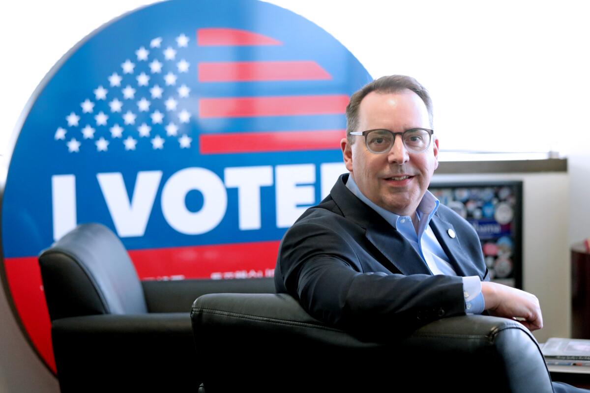 Dean Logan, in glasses and business suit, smiles in front of an "I Voted" sign.