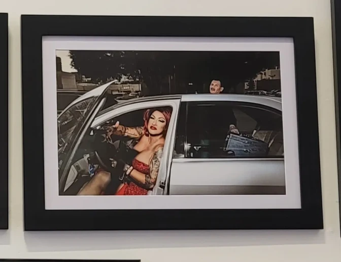 A framed photograph on a wall of a female-presenting person, wearing a red dress and has tattoos, in the drivers seat with the door open and another person right outside the right backseat passengers door.