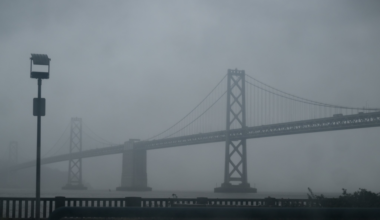 A large suspension bridge is seen through thick fog, with a railing and a tall lamp post in the foreground.