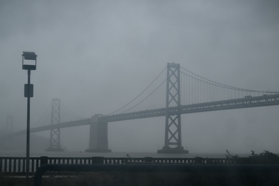 A large suspension bridge is seen through thick fog, with a railing and a tall lamp post in the foreground.