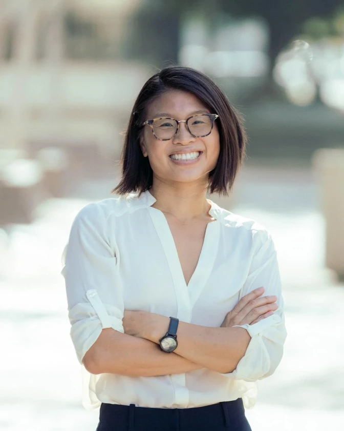 A woman with glasses in a white blouse stands with her arms folded smiling at the camera.