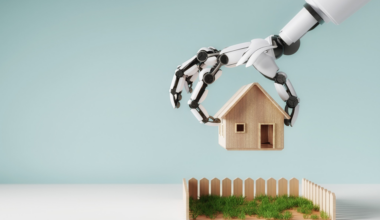 A robotic hand is holding a small wooden house above a fenced patch of grass against a light blue background.