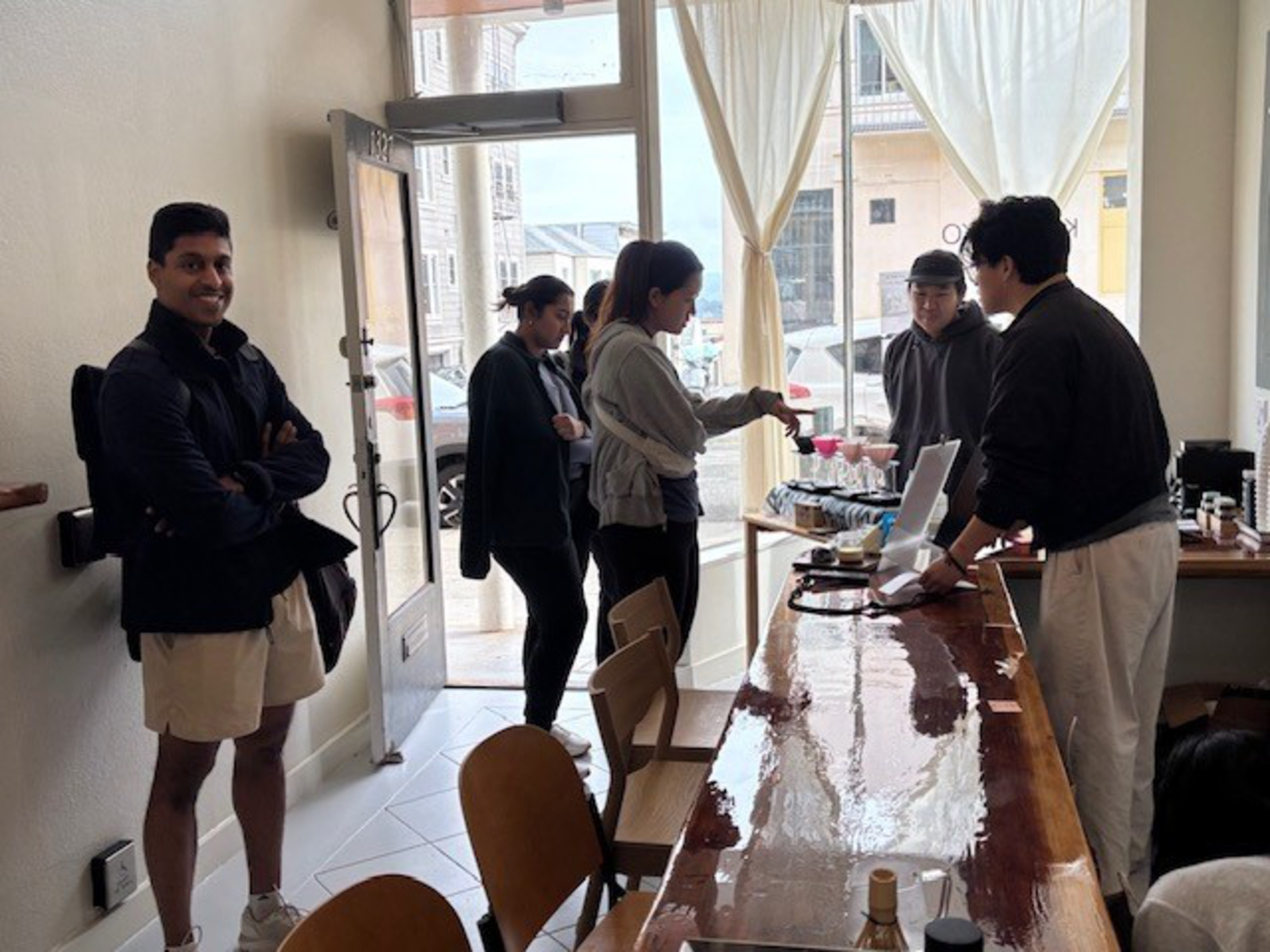Five people interact inside a bright café, where a man behind a wooden counter serves three customers as another man stands smiling near the open door.