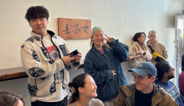 A group of people socialize indoors near a wooden sign with Chinese characters, with some smiling, one person flashing a peace sign, and others engaged in conversation.