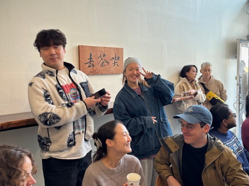 A group of people socialize indoors near a wooden sign with Chinese characters, with some smiling, one person flashing a peace sign, and others engaged in conversation.