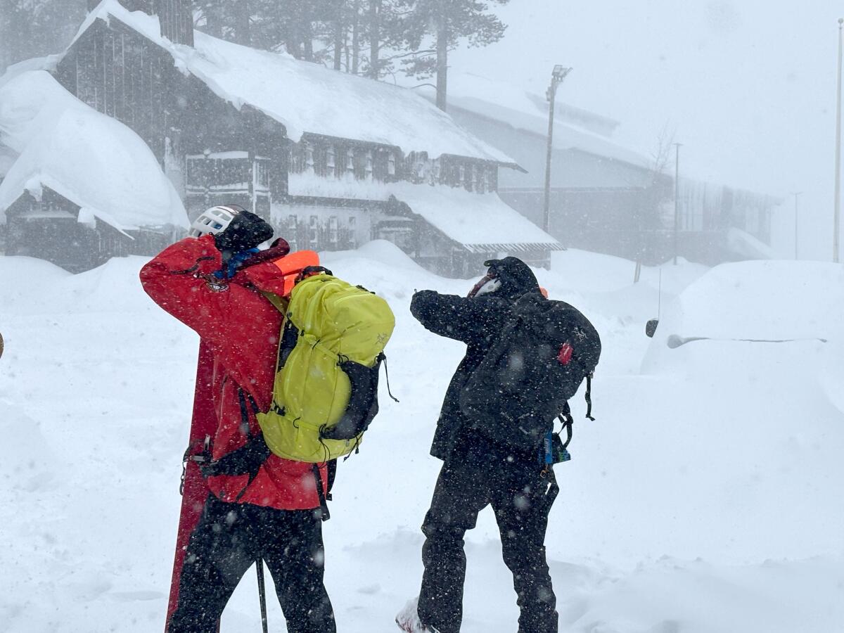 Two people in snow clothing wearing backpacks walk in snow 