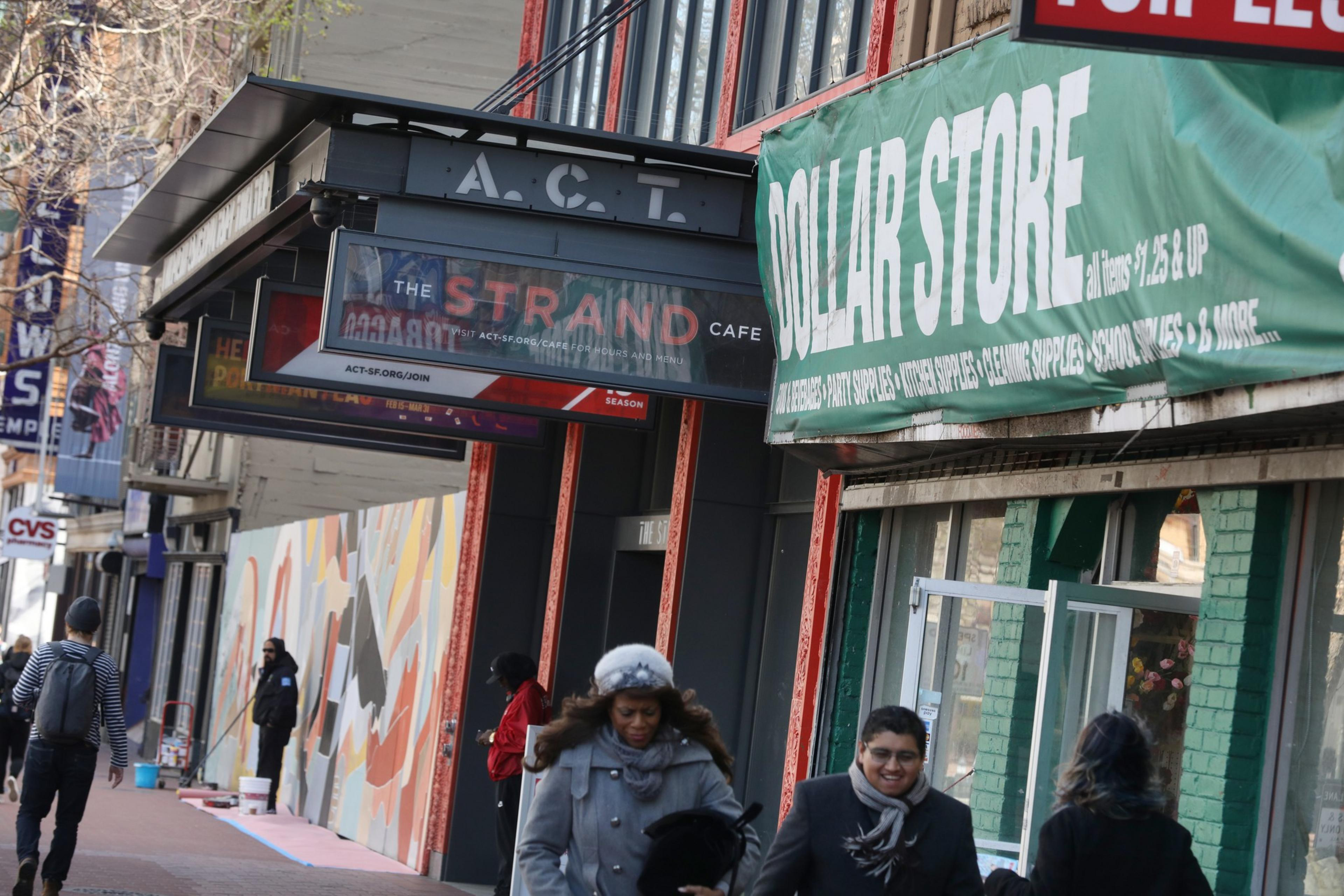 A busy sidewalk with people walking past a dollar store and The Strand Cafe, with colorful murals and other storefronts visible down the street.