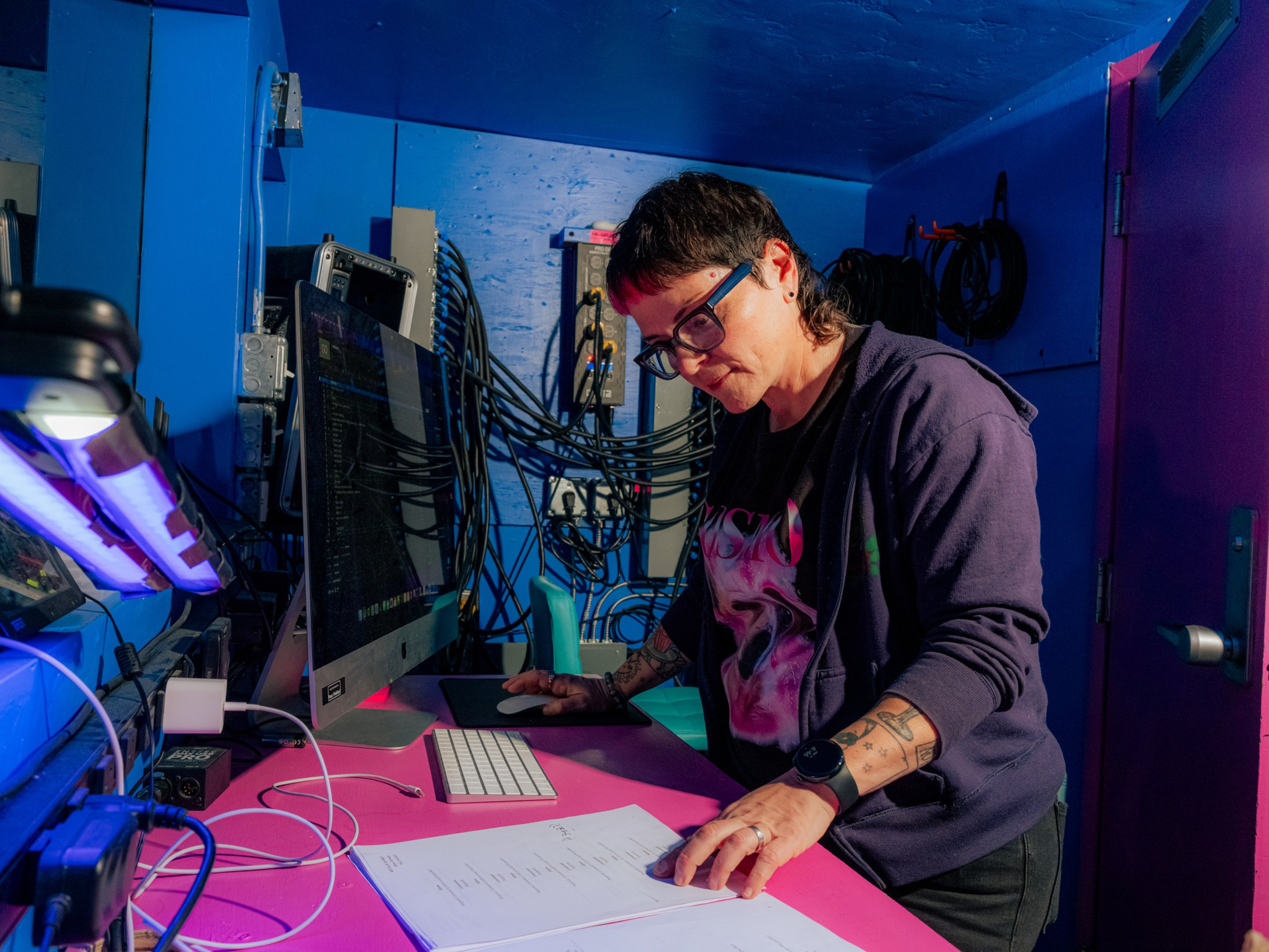 A person with glasses and tattoos is working at a pink desk with a computer, reviewing documents in a room with blue walls and exposed cables.