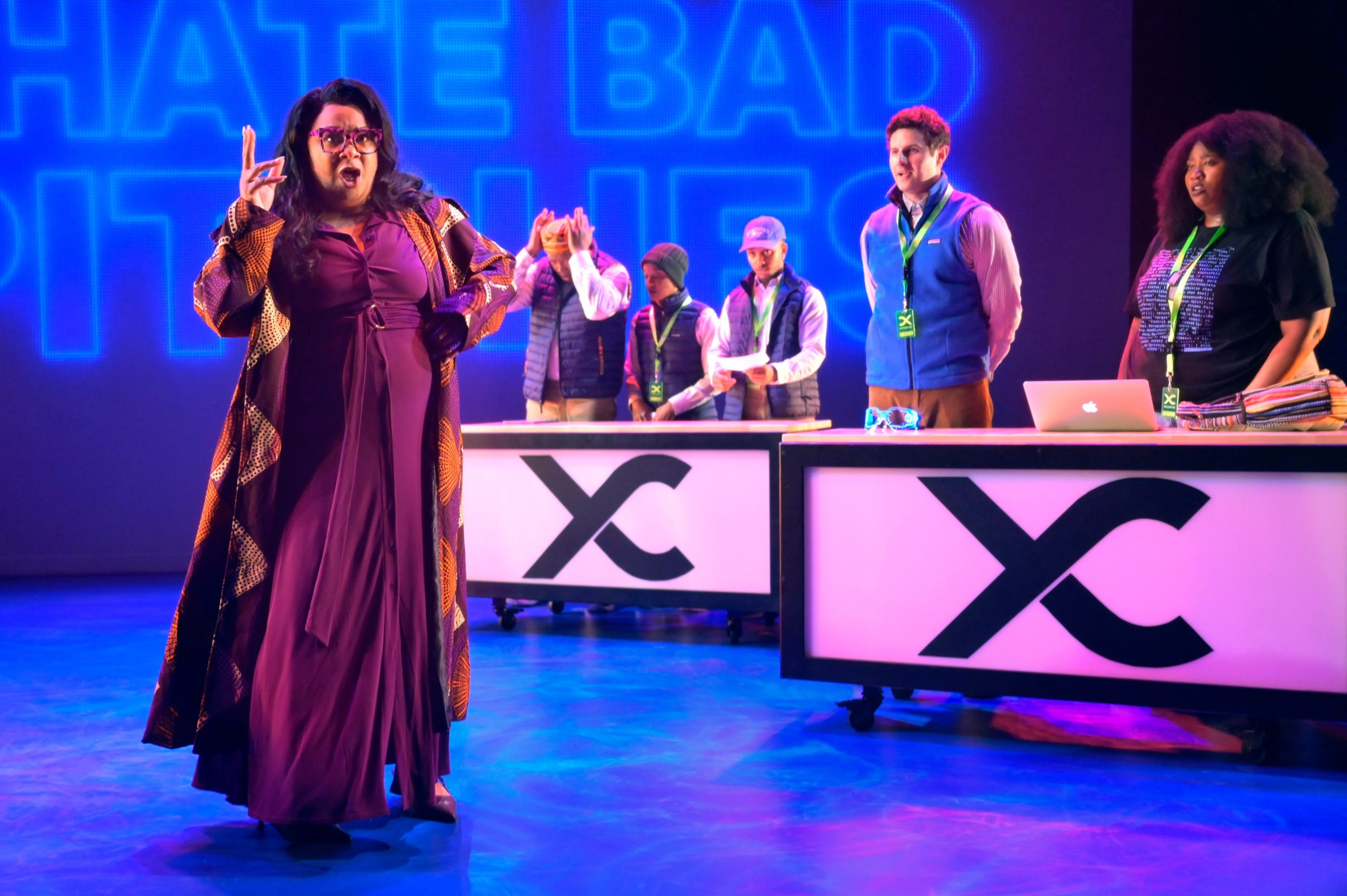 A woman in a long dress and patterned coat gestures dramatically while five people stand behind illuminated desks with “X” logos and wear lanyards.