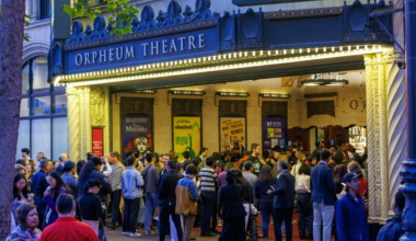 A large crowd gathers outside the illuminated Orpheum Theatre entrance with show posters visible inside.
