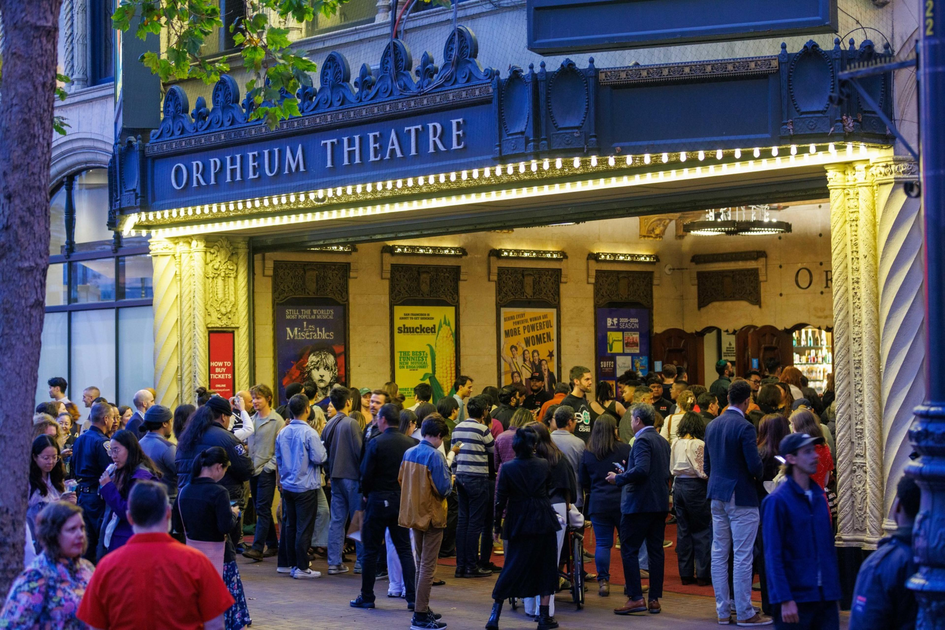 A large crowd gathers outside the illuminated Orpheum Theatre entrance with show posters visible inside.