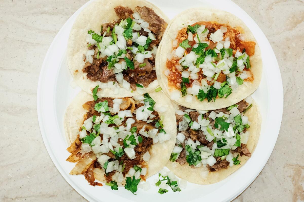 Four tacos in soft corn tortillas covered in onions and cilantro on a white plate