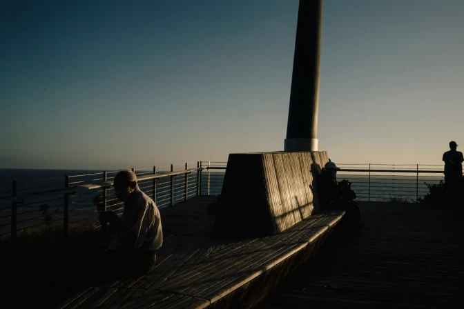 A man, and other people in the background, sit at an area overlooking the ocean. They are all partially lit by the sun at sunset.