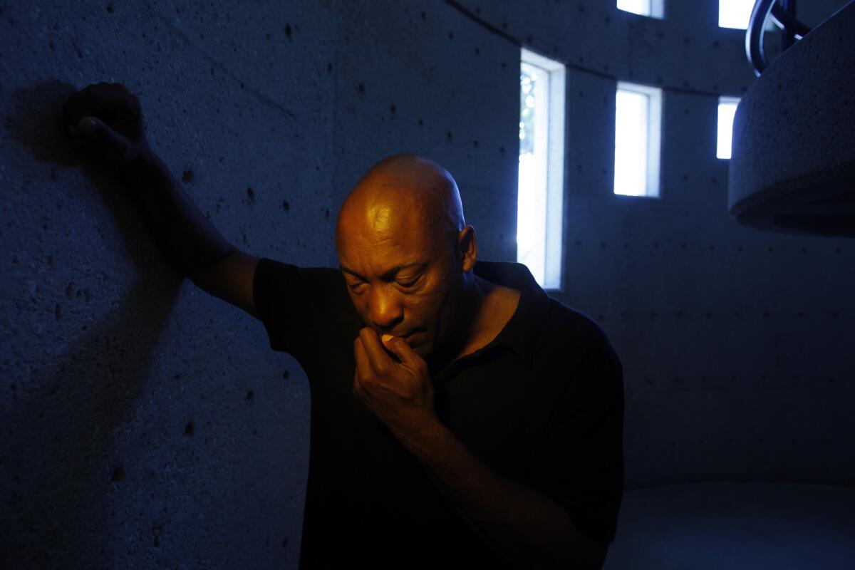 A man with a shaved head, in a dark shirt, poses for a photo while leaning against a wall