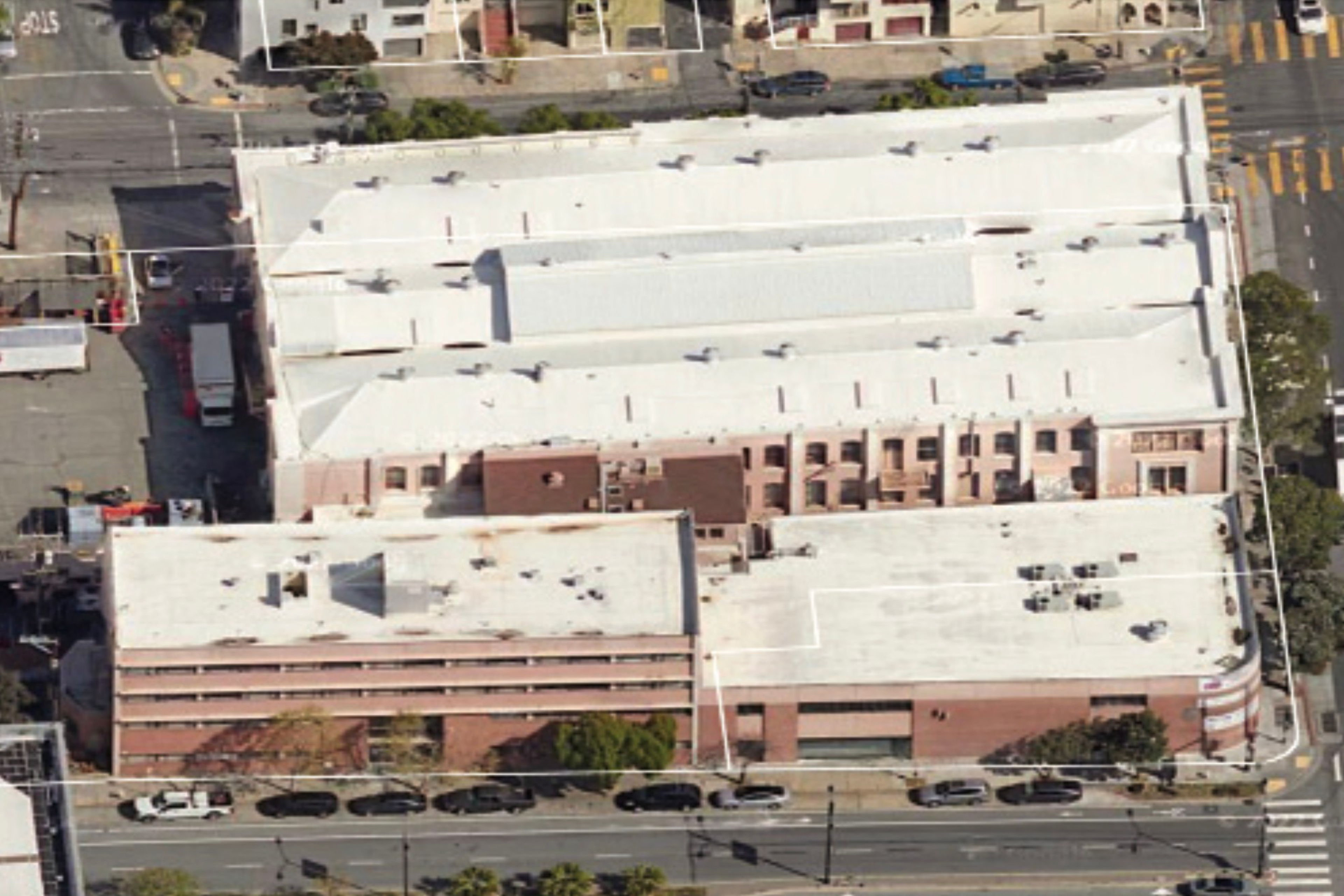 Aerial view of a large, rectangular building with white roofs, surrounded by streets on all sides and parked cars along the roads.