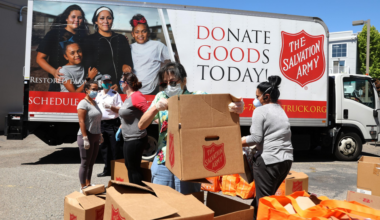 Volunteers wearing masks and gloves unload and organize Salvation Army donation boxes and orange bags near a donation truck on a sunny day.