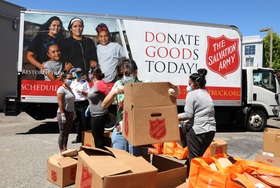 Volunteers wearing masks and gloves unload and organize Salvation Army donation boxes and orange bags near a donation truck on a sunny day.