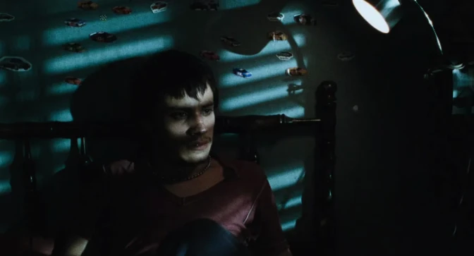 A man with medium skin tone sits in a bed in a dark room with light coming in slats through the blinds. There are car stickers on the wall.