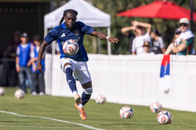 A Black man in a blue soccer uniform juggles a ball near the sideline.