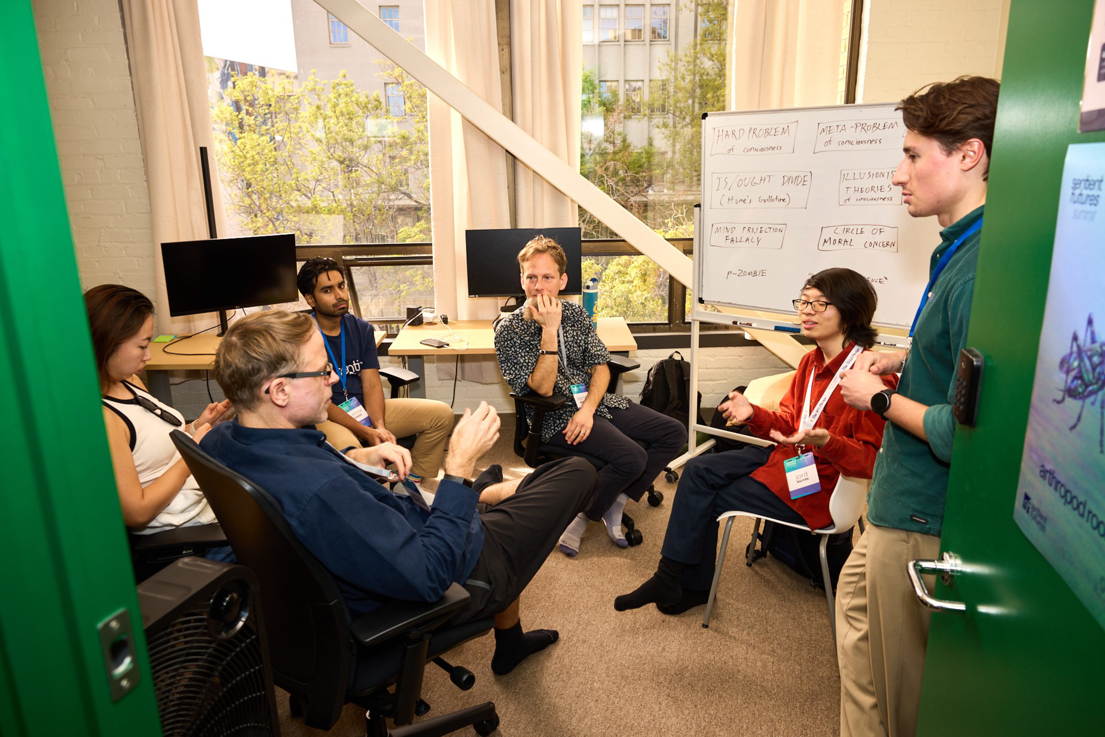 Five people sit and stand in an office room engaged in discussion near two monitors and a whiteboard with notes about problems and theories.