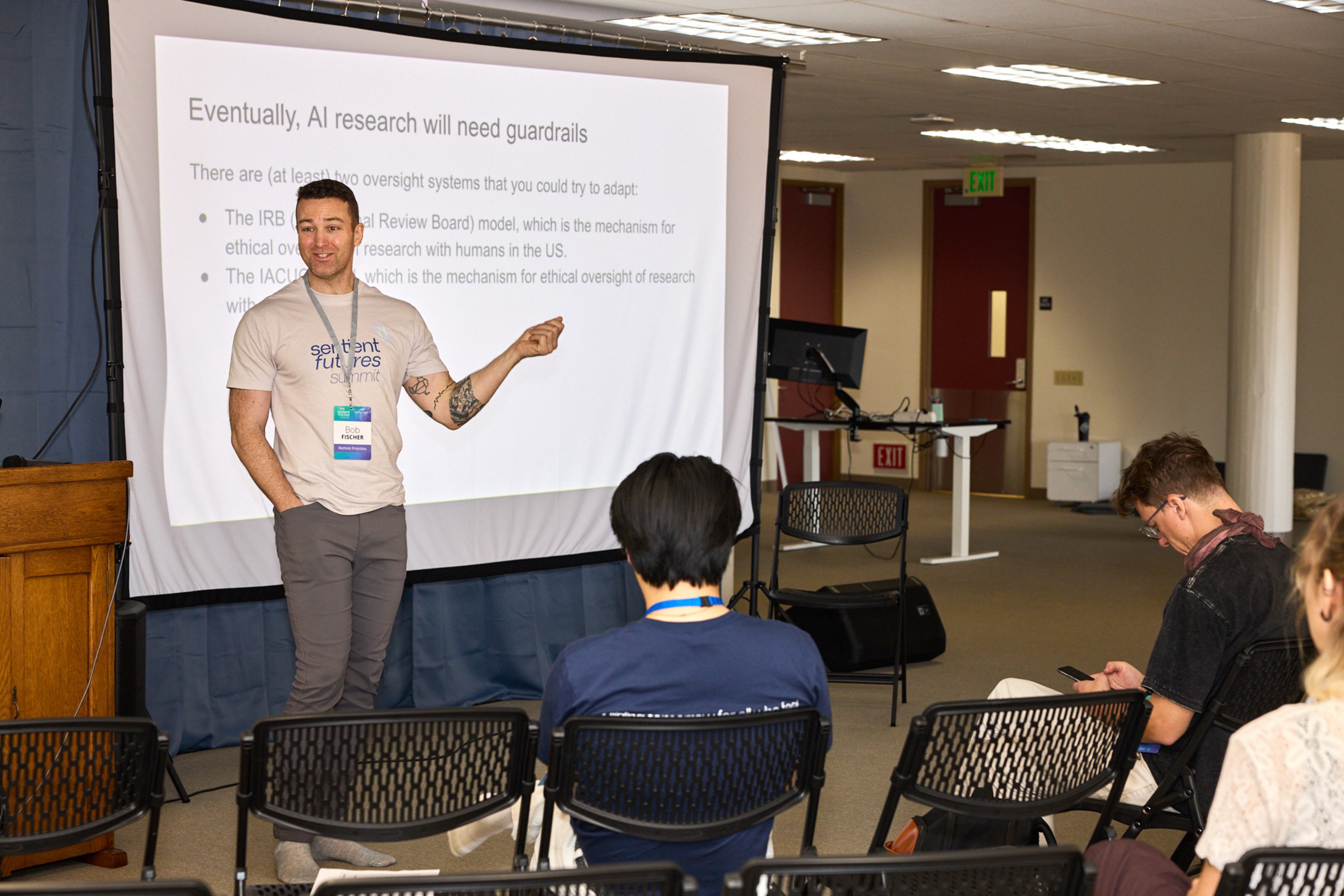 A man wearing a “Sentient Futures Summit” shirt is presenting about AI research guardrails to a small seated audience, with a projection slide behind him.