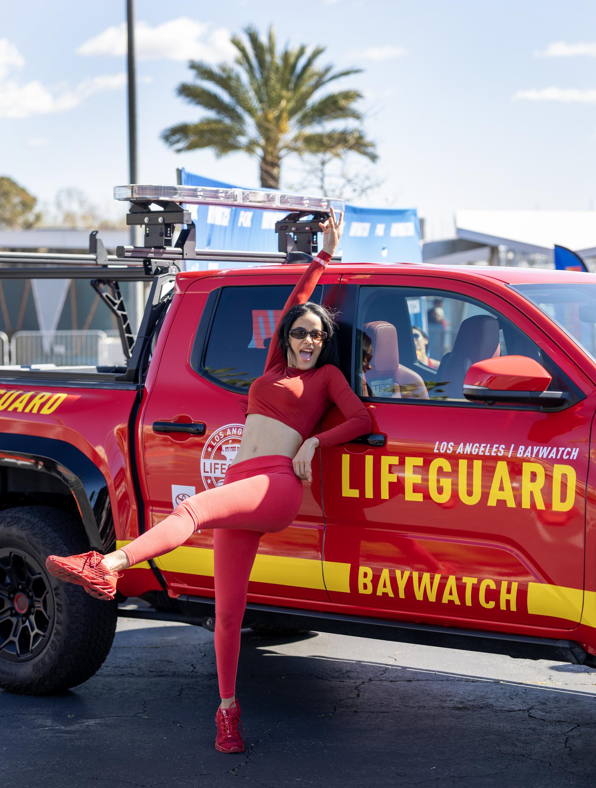 A woman in a red top and leggings leans against a red truck with her hand and leg in the air.