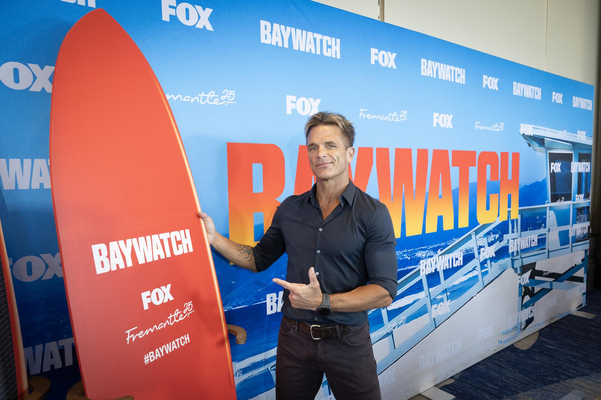 A man in a dark shirt and pants points at a Baywatch surfboard.