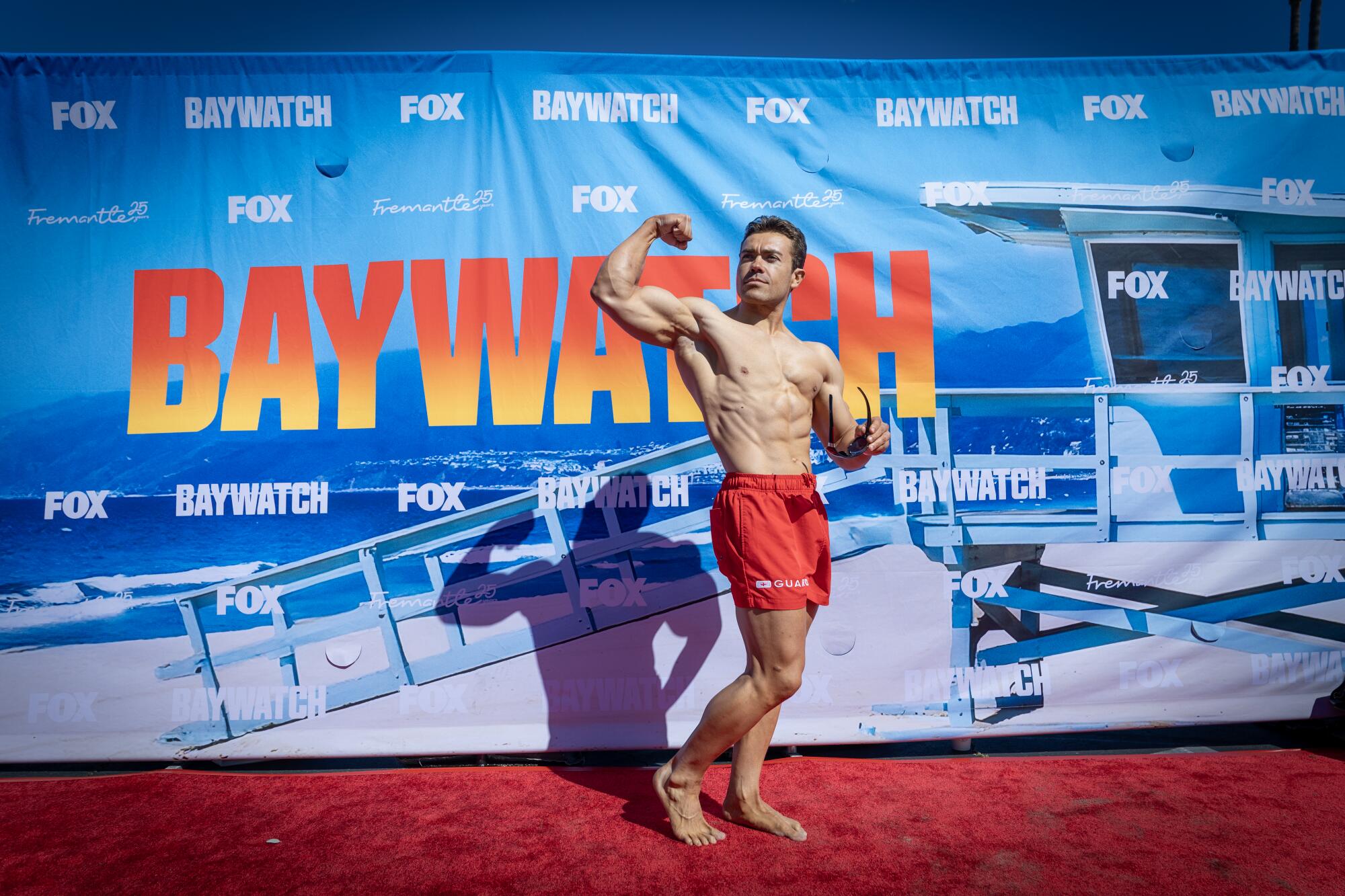 A muscular man flexing on a red carpet with a banner that says Baywatch across it.