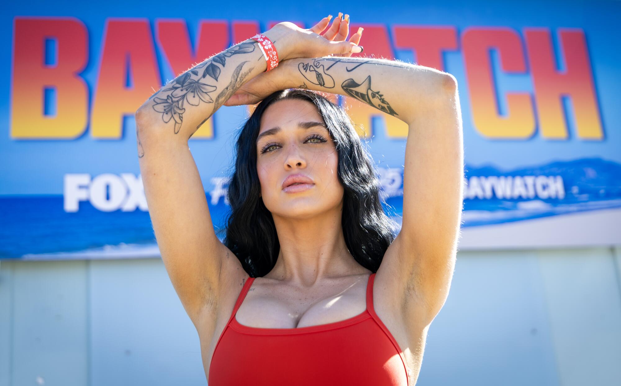 A woman in red swimsuit lifts her tattooed arms above her head.